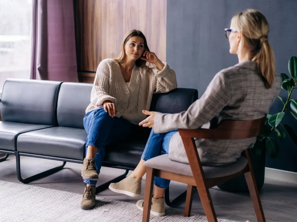 A young woman sits on a modern black leather couch, attentively listening to a professional therapist in a private consultation room, representing psychological assessment services in Los Angeles.