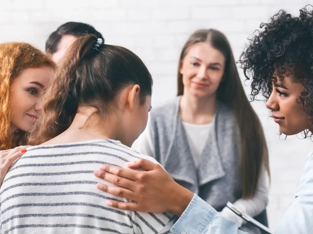 A supportive therapy group session where a young woman receives comforting gestures from fellow members, creating an atmosphere of empathy and understanding.