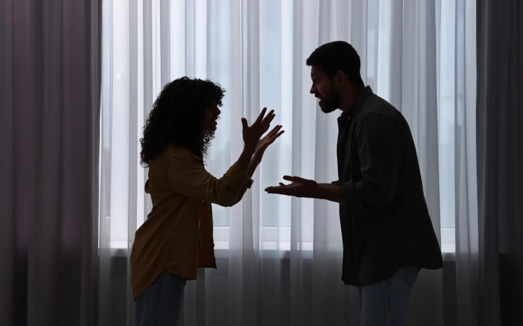 A couple arguing in a darkened room in front of drawn net curtains