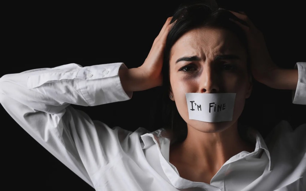 A stressed woman with her hands on her head pulling her hair out with a sticker over her mouth that reads "I'm fine", symbolizing the impact of domestic violence on the Victim
