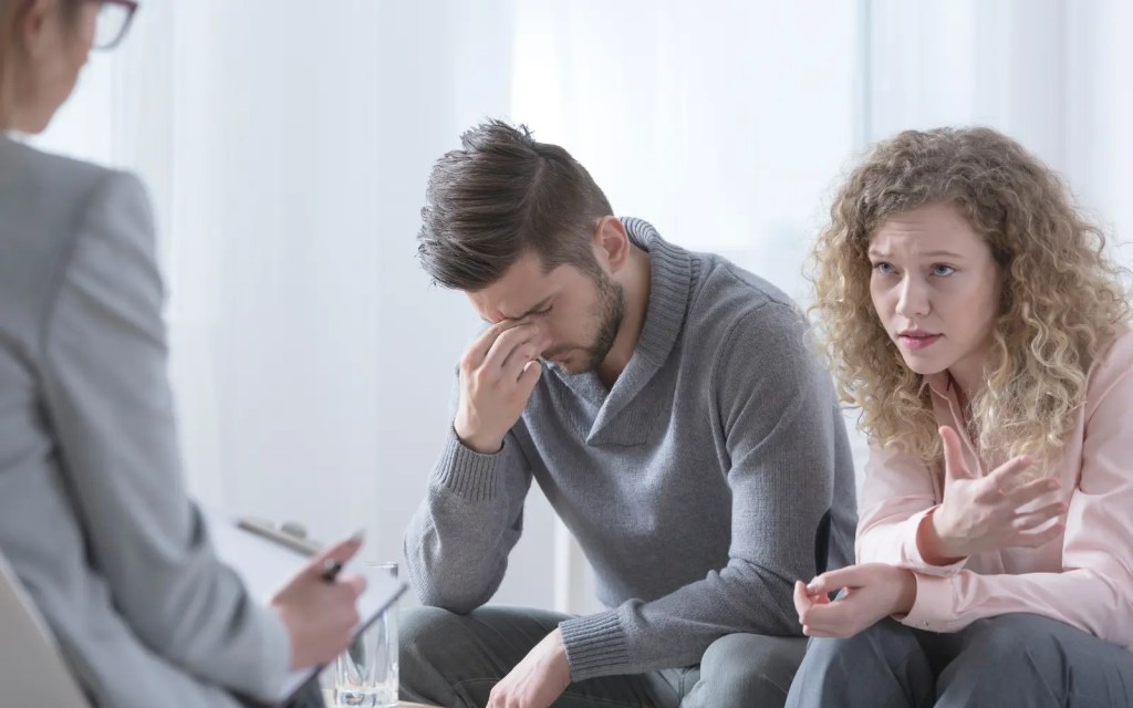 A man and woman in a couple therapy session, the man is holding his nose and looking down because of the fear of couples therapy