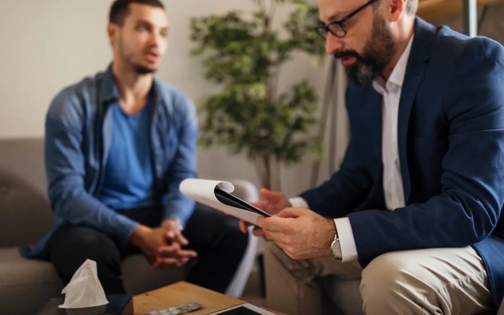 A man in an individual therapy session with a male therapist sitting on a couch while the therapist makes notes on his clipboard