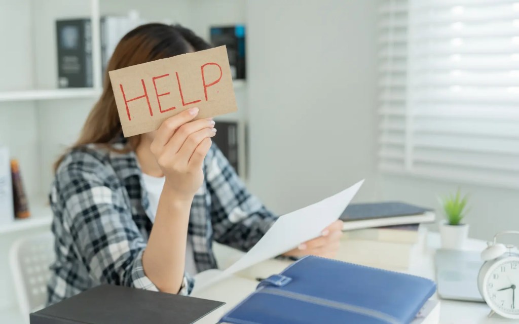 A woman sitting at a desk holding up a piece of paper with the word help written on it representing relationship difficulties experience by neurodivergent to couples