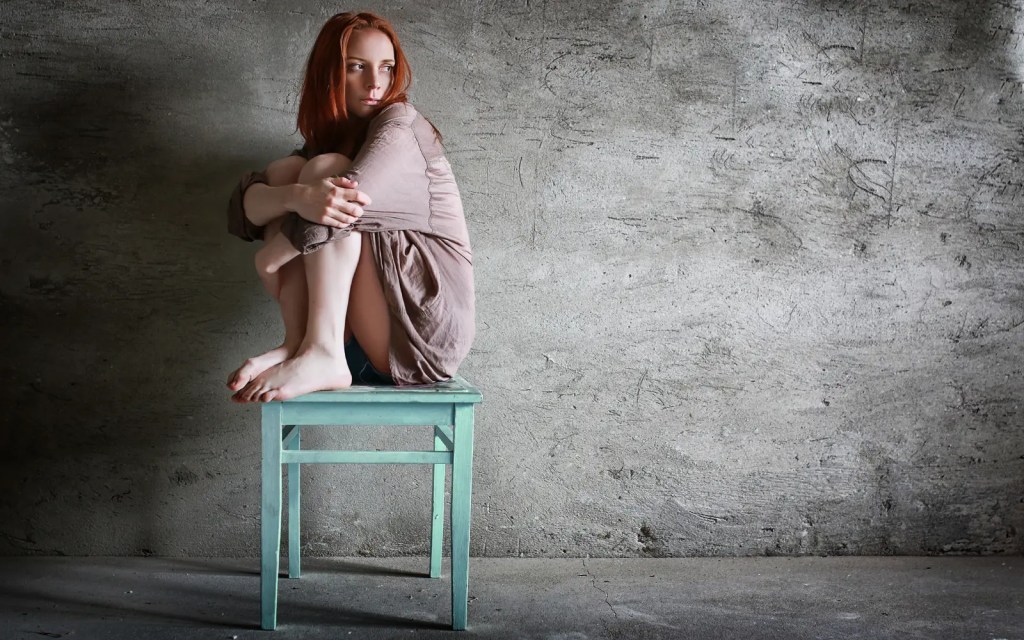 A woman in front of a gray wall sitting on a dirty wooden chair with her knees pulled up to her chest and her arms wrapped around her legs, symbolizing the impact of trauma in relationships