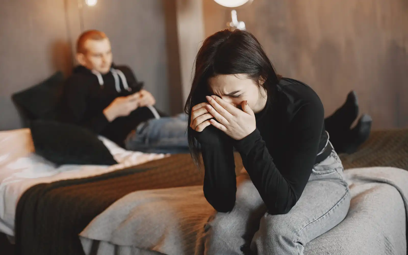 A woman sitting on the edge of the bed crying into her hands while eyes on the bed behind her looking at his phone. The woman has unresolved trauma that could be dealt with in either individual or couples therapy.