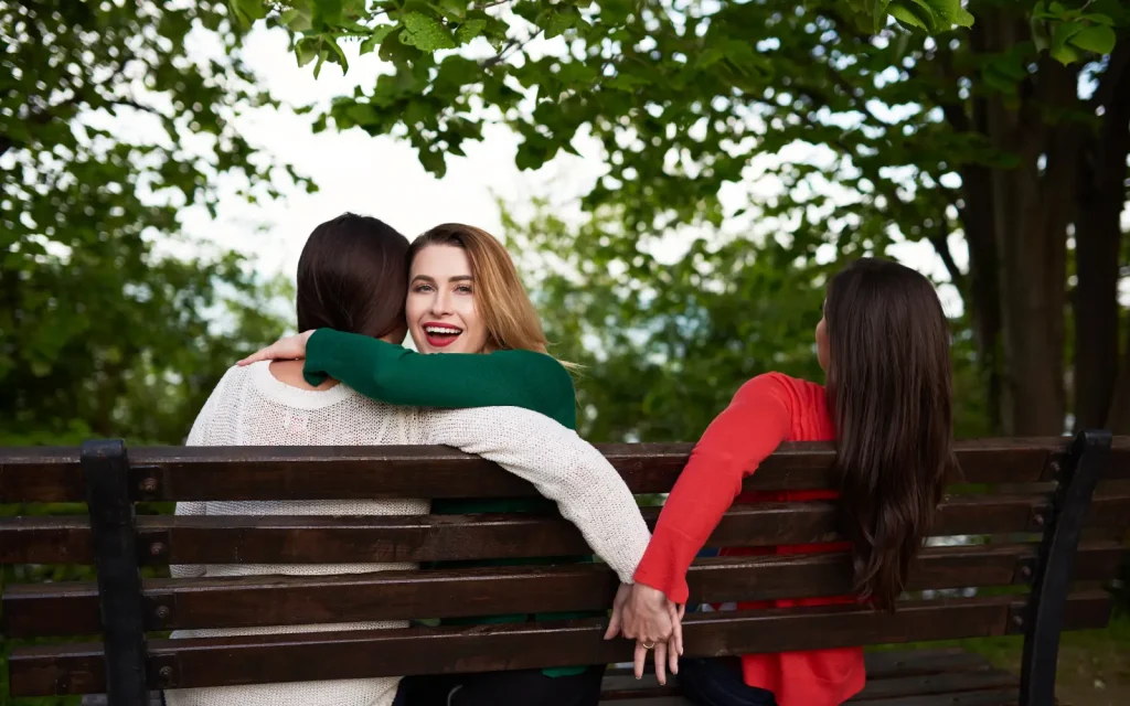 Three women on a bench, two embracing and the third covertly holding hands with one of the other two, representing infidelity in relationships