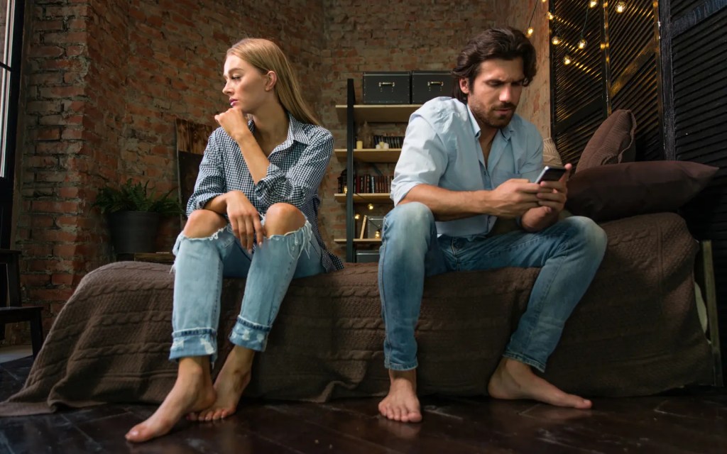 A couple sitting on a bed, looking away from each other with the man on his phone, representing unhealthy dynamics in relationships