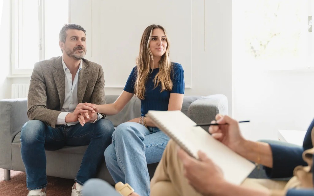 A couple sitting on a sofa in a couples therapy session, holding hands and looking at the therapist with hope, representing the importance of attending weekly couples therapy sessions for the best progress