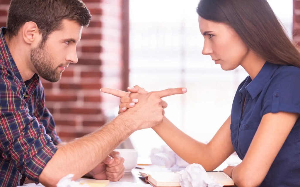 A heterosexual couple facing each other across a table and pointing fingers at each other, representing couples getting defensive in couples therapy