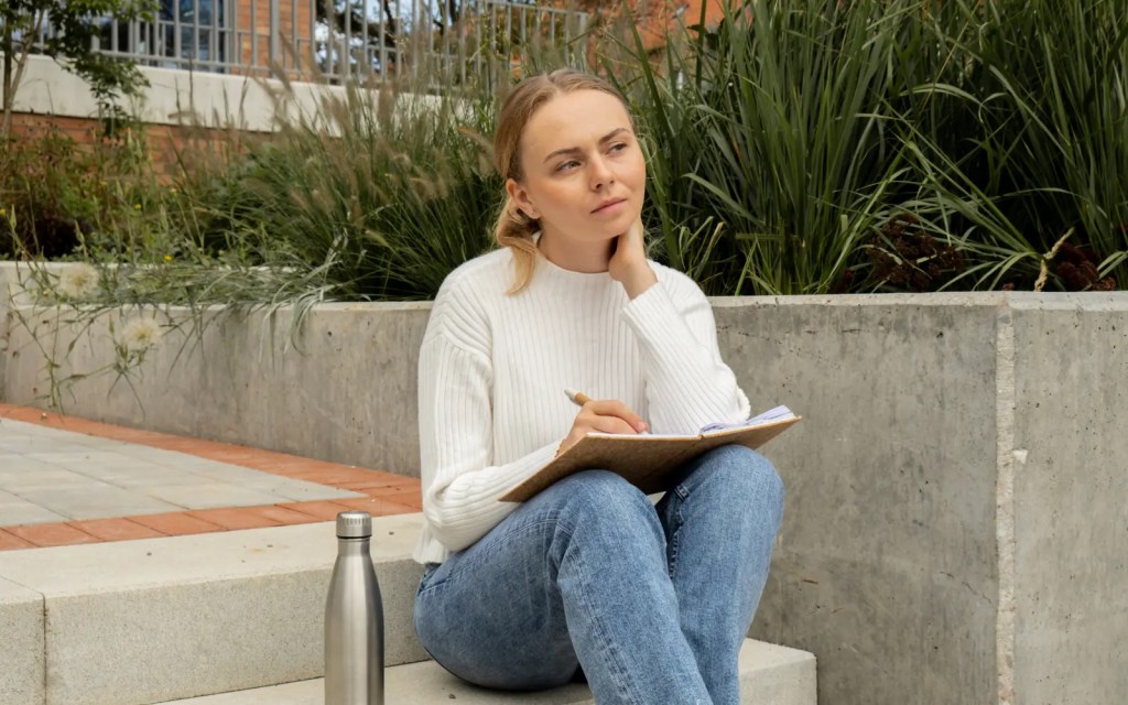 A woman sitting on steps outside looking pensive and holding a journal, representing the self-reflection that's possible through therapy