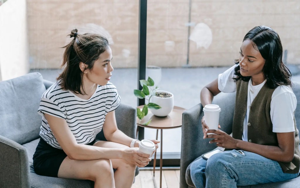 Two women sitting opposite each other holding coffee, looking at each other intently while they are discussing difficult topics after a marriage counseling session.