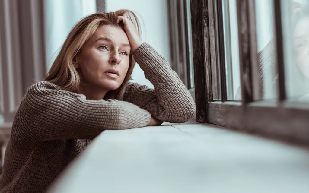 Safety-in-couples-therapy-Los-Angeles A woman looking out of a window with her head in her hand in despair, representing the importance of safety in couples therapy